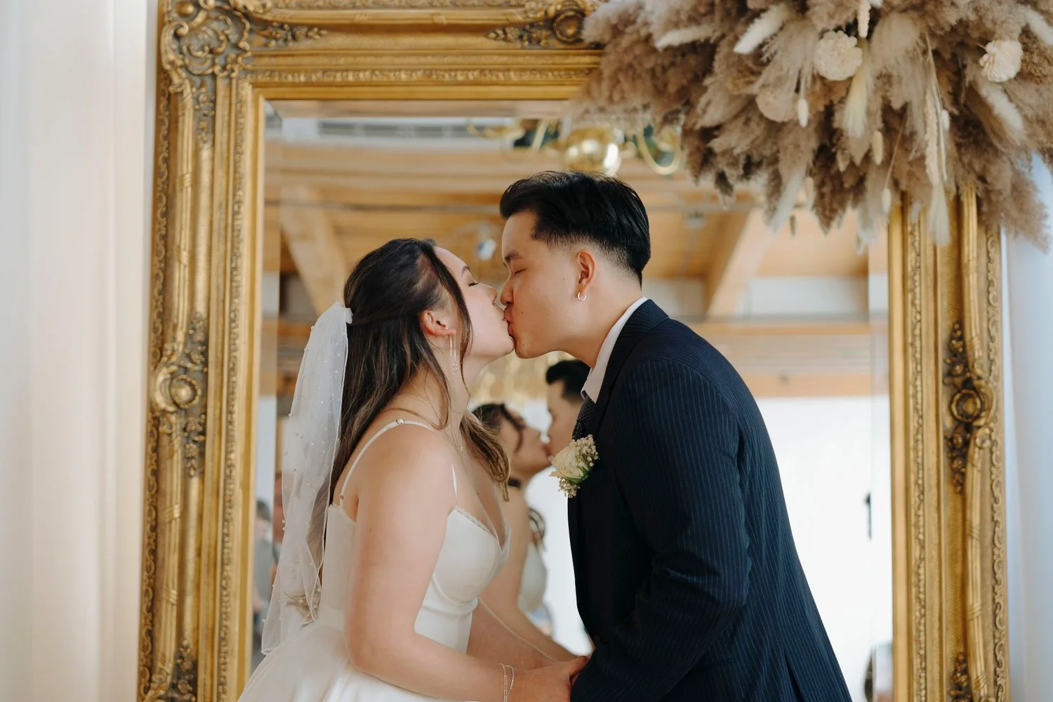 Couple in wedding attire kissing in front of gilded mirror with floral arrangement above.