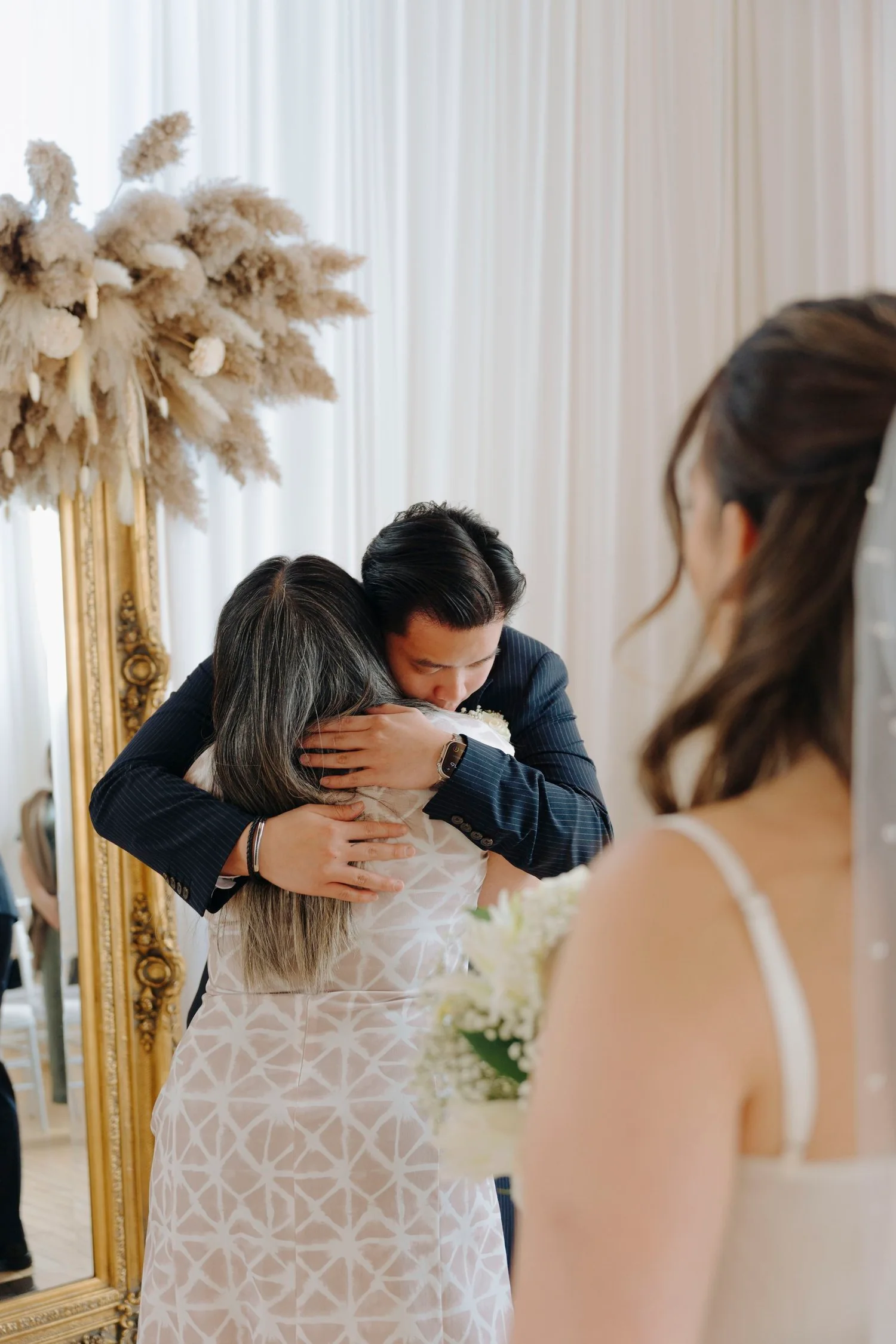 A man in a navy suit hugging a woman with gray hair, with another woman holding a bouquet in the foreground at a wedding celebration.