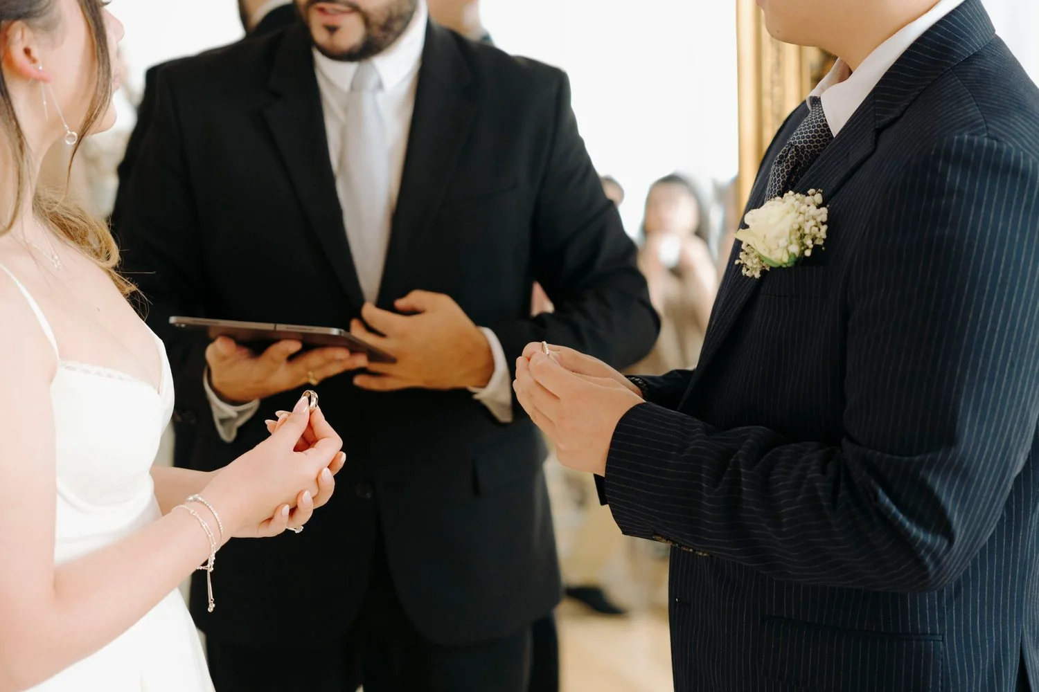 A couple getting married exchanging rings during their wedding ceremony with officiant holding a tablet in the background.