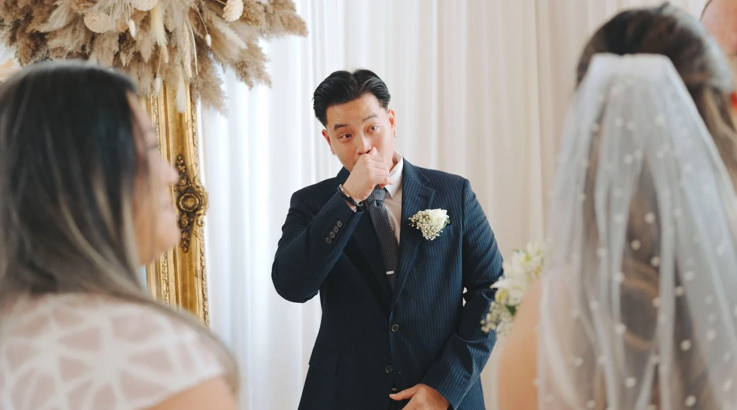 A groom dressed in a dark pinstripe suit with a white flower boutonniere, wiping his nose or covering his mouth with his hand. The bride, partially visible in a white veil, and two women with blurred features are in the foreground. The setting is decorated with white and gold, possibly for a wedding ceremony.