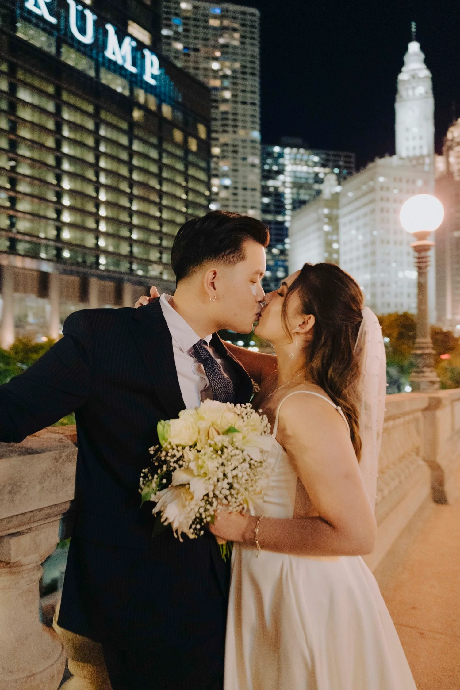 A newlywed couple sharing a kiss at night on a city bridge, with tall buildings and illuminated signage in the background.