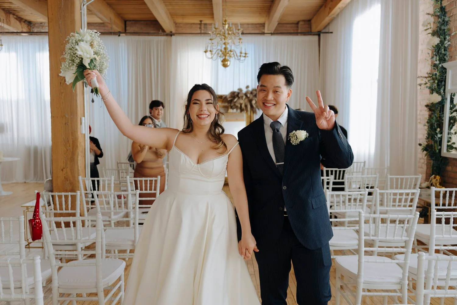A bride and groom celebrating at their wedding ceremony, smiling, holding hands, with the bride raising a bouquet, inside a decorated venue.