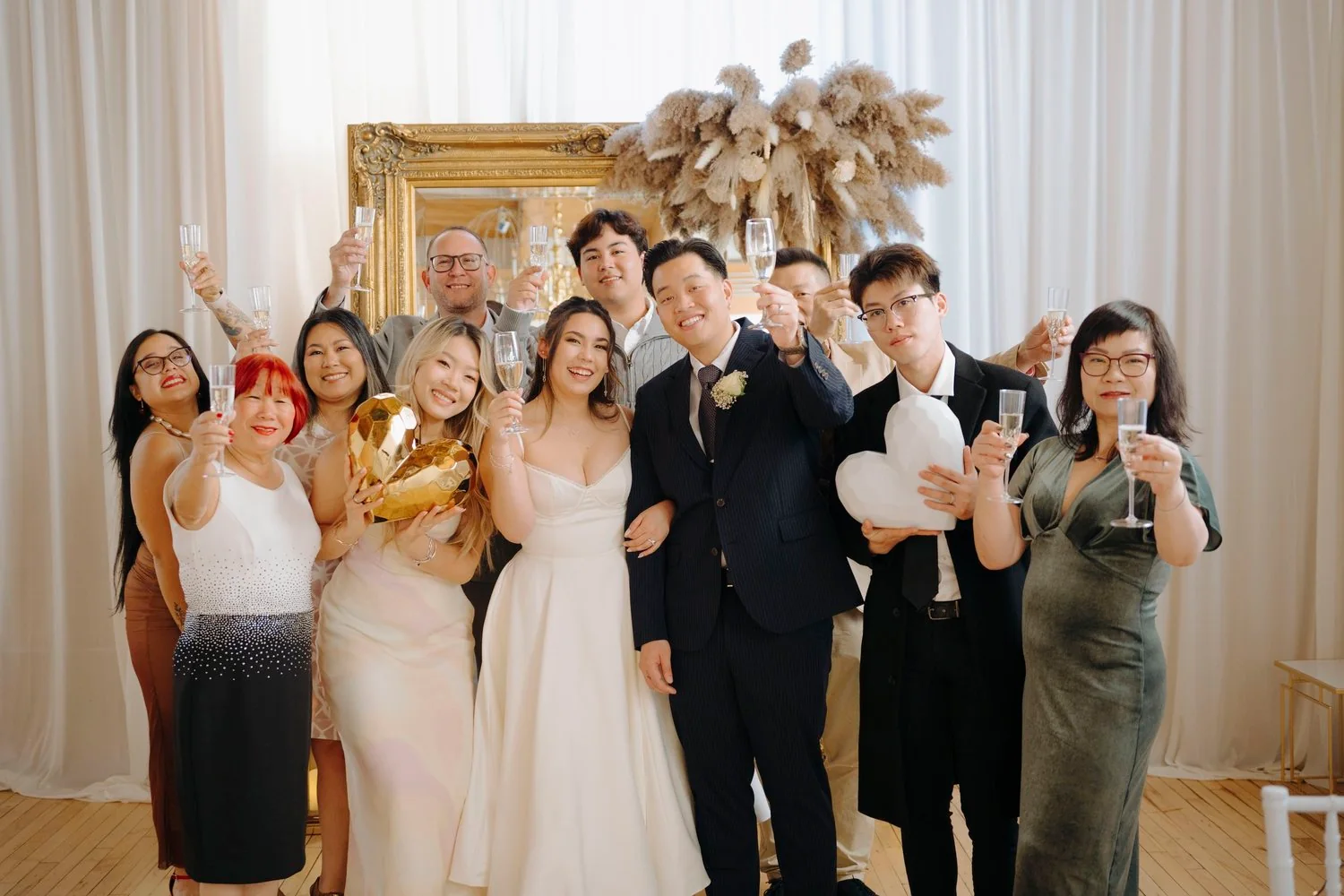 Group of people at a wedding celebration raising champagne glasses, smiling, in elegant attire with a bride and groom in center, in a decorated venue.