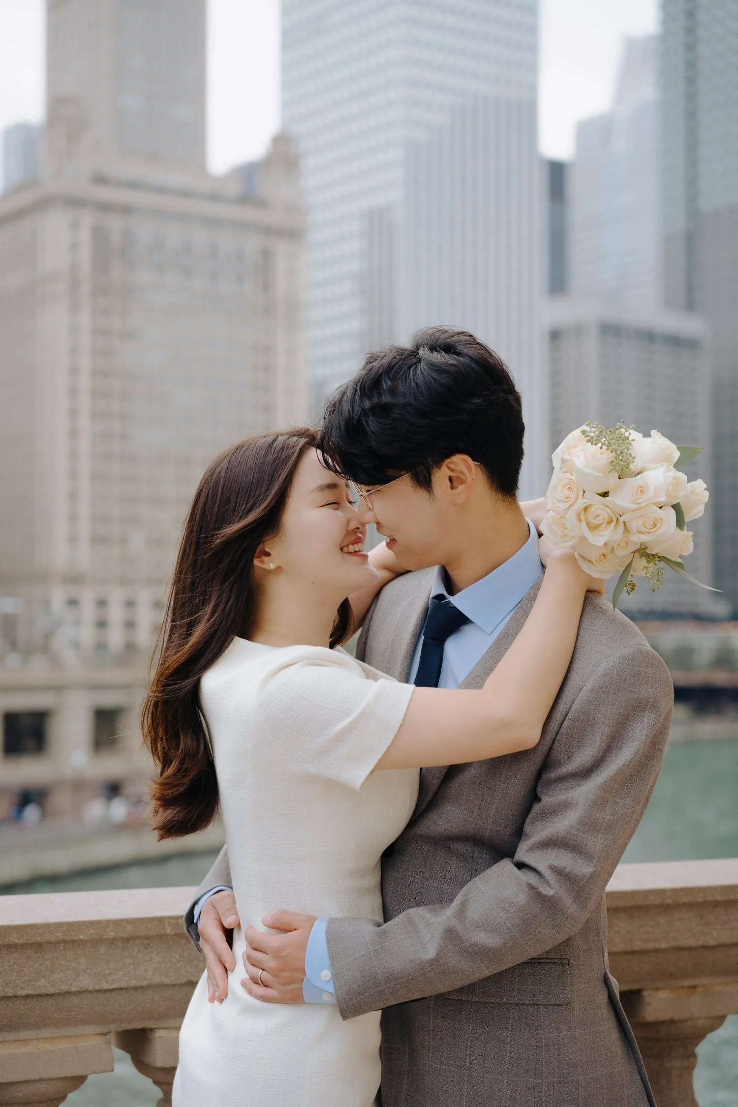 A happy couple embracing on a bridge in a city with tall buildings, the woman holding a bouquet of white roses.