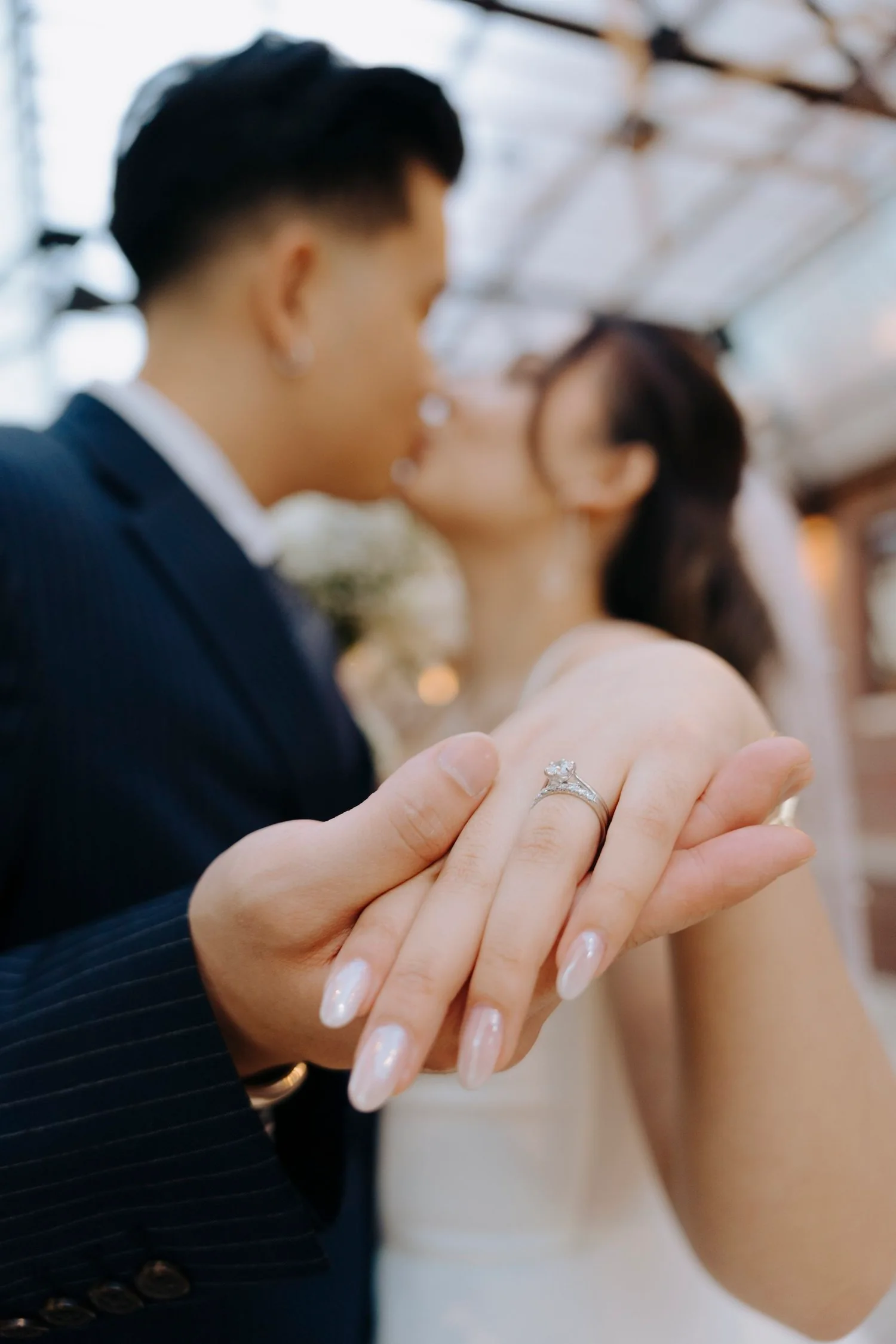 Close-up of a woman's hand showing an engagement ring, with a couple kissing blurred in the background.
