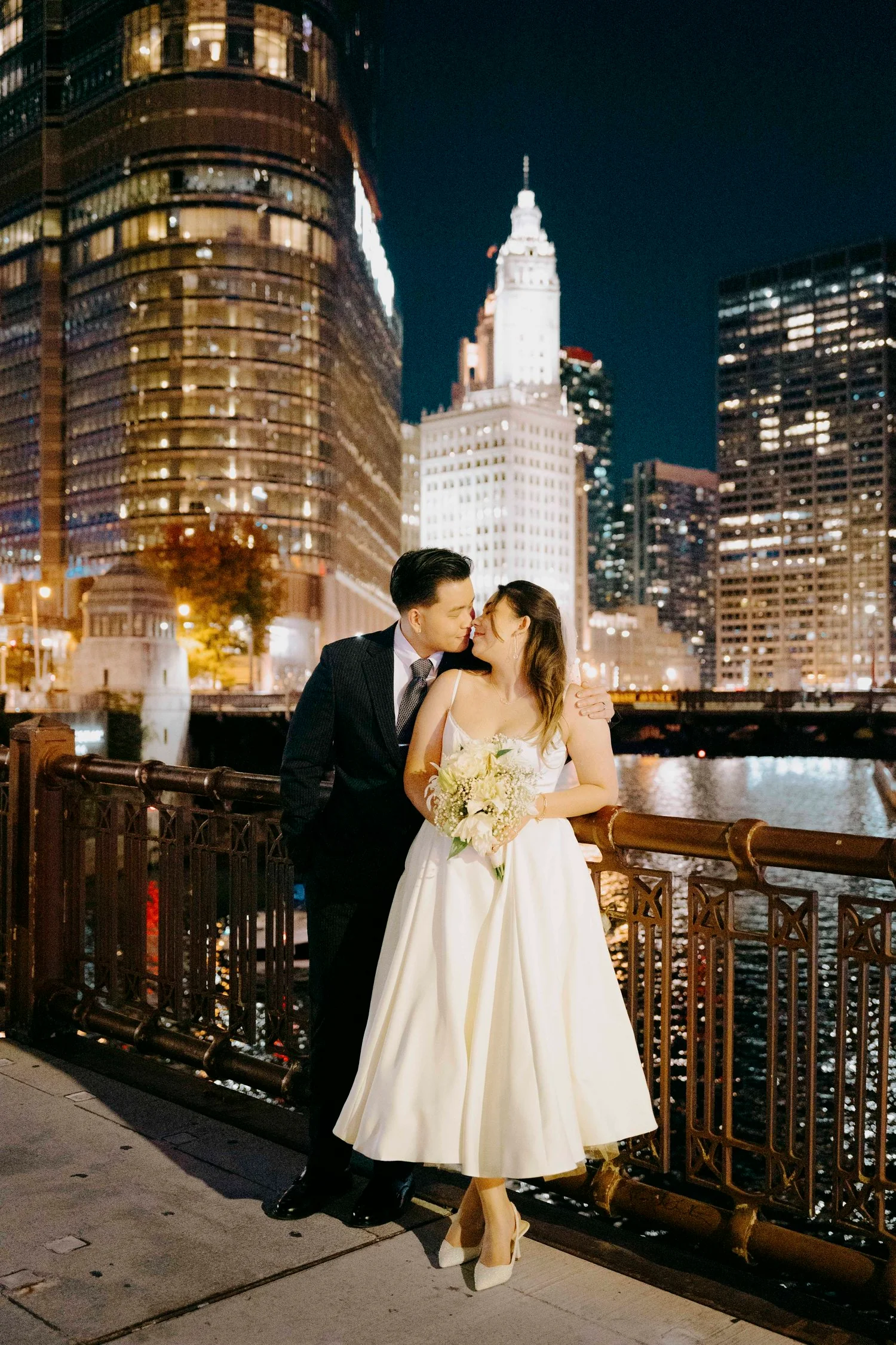 A newlywed couple sharing an intimate moment on a riverside promenade at night, with the Manhattan skyline lit up in the background, including the Empire State Building.