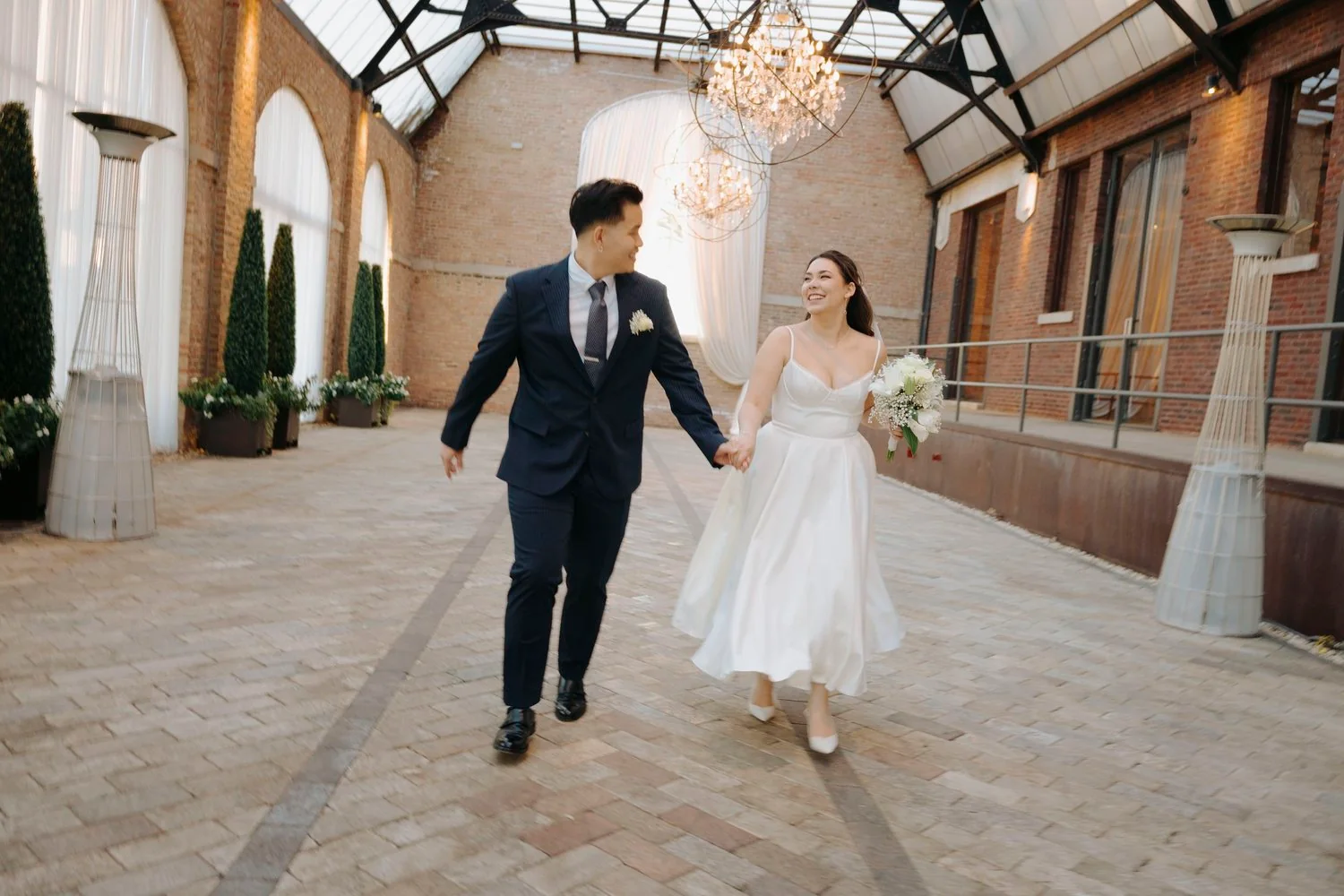 Bride and groom holding hands and smiling while walking inside a decorated venue with brick walls, large windows, and chandeliers.