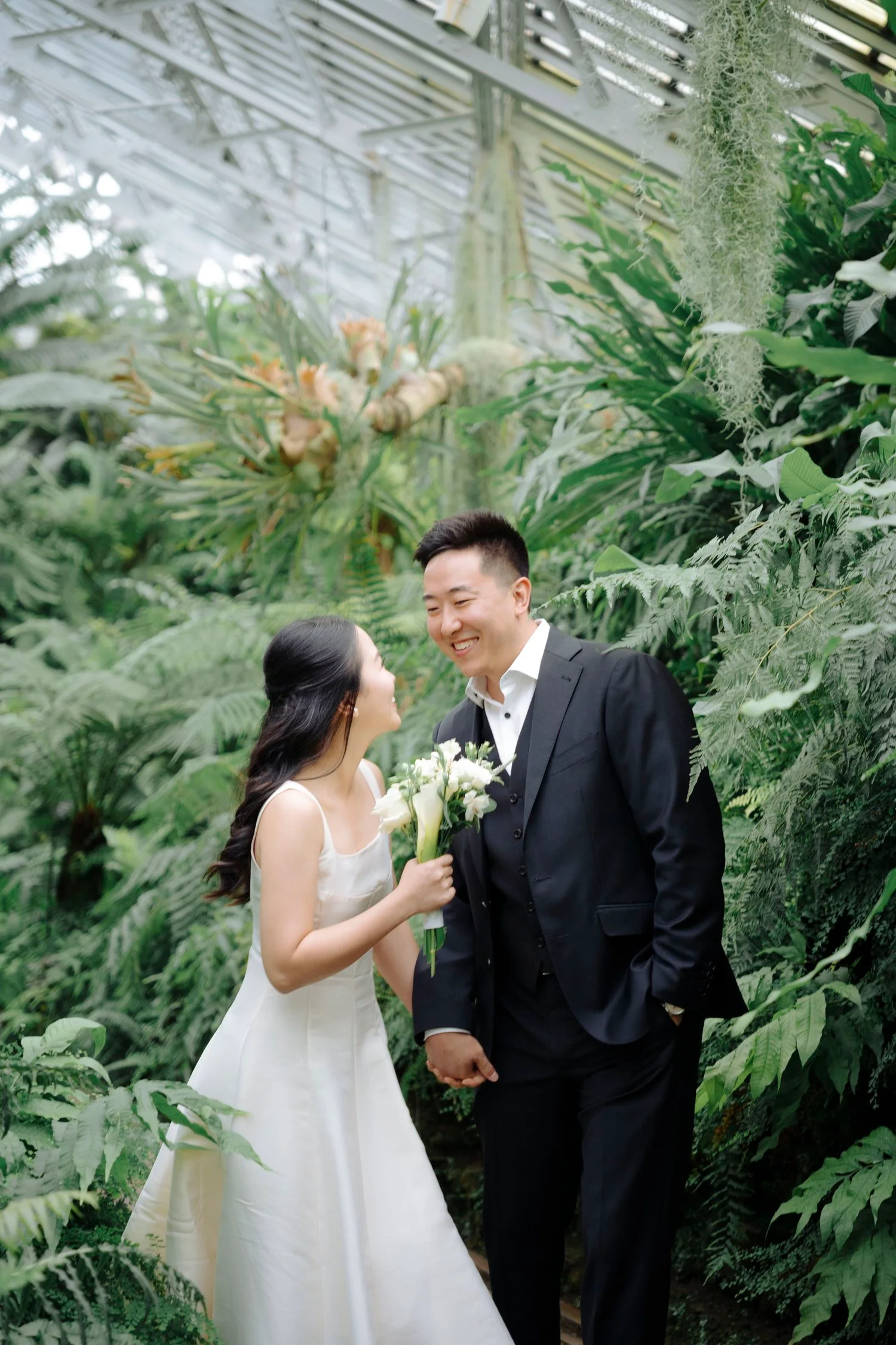 A bride and groom smiling and holding hands inside a lush, green greenhouse. The bride is holding a bouquet of white flowers, and they are looking at each other happily.