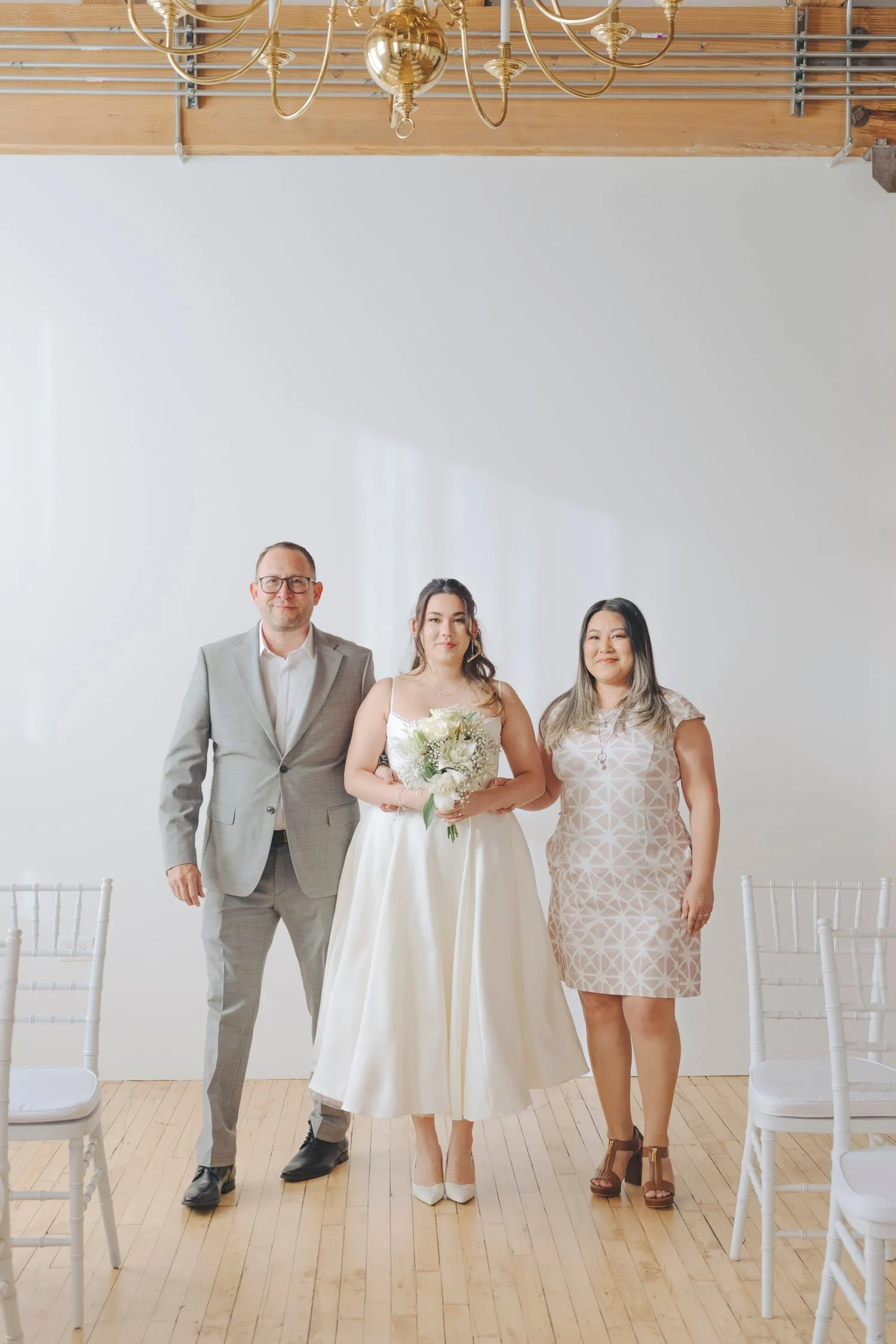 A bride standing between a man in a gray suit and a woman in a patterned dress, all smiling and posing indoors with white chairs and a chandelier overhead.