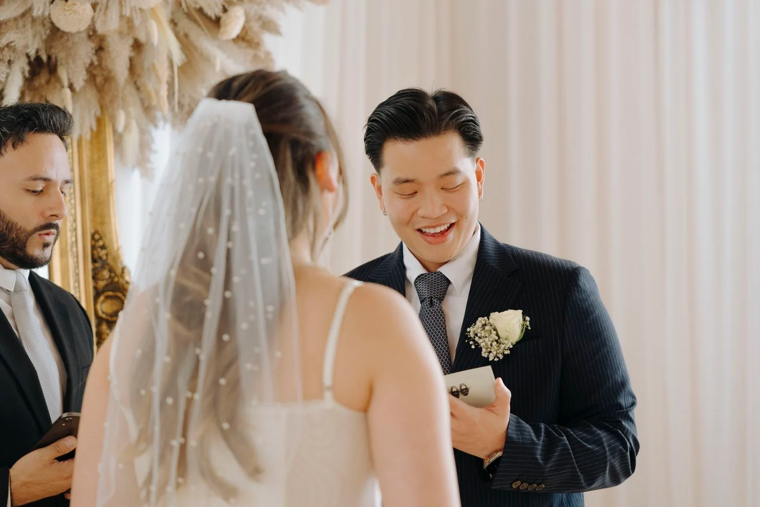 A wedding ceremony with a groom smiling while reading vows to the bride, who is wearing a veil. A man on the left looks at a phone. The setting is elegant with cream-colored curtains and ornate decorations.