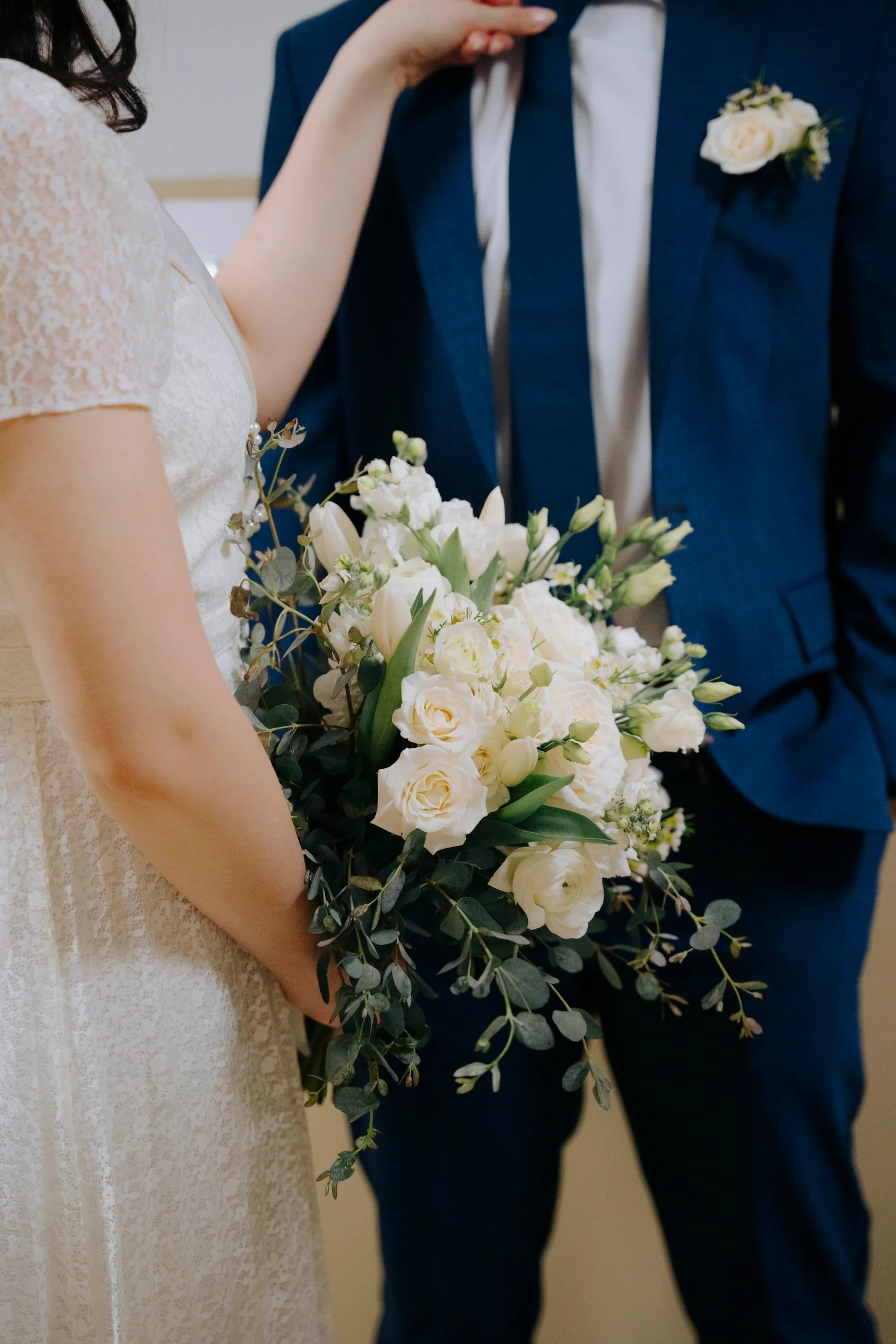 A bride holding a bouquet of white roses and greenery, standing next to a groom in a navy suit with a white boutonniere.