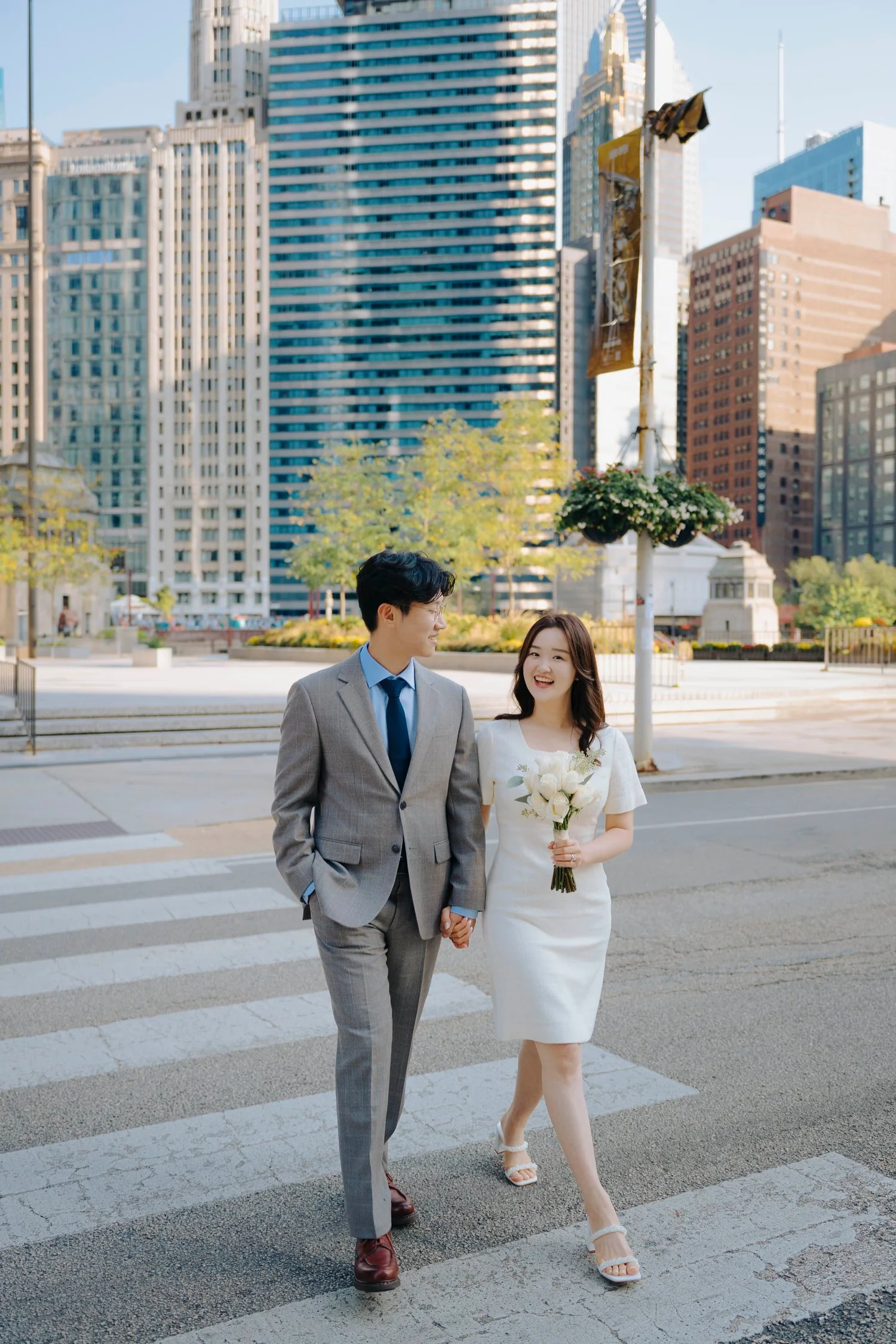 A newlywed couple holding hands and walking across a crosswalk in a city with tall buildings, the woman holding a bouquet of white roses and wearing a white dress, and the man in a gray suit with a blue tie, both smiling.