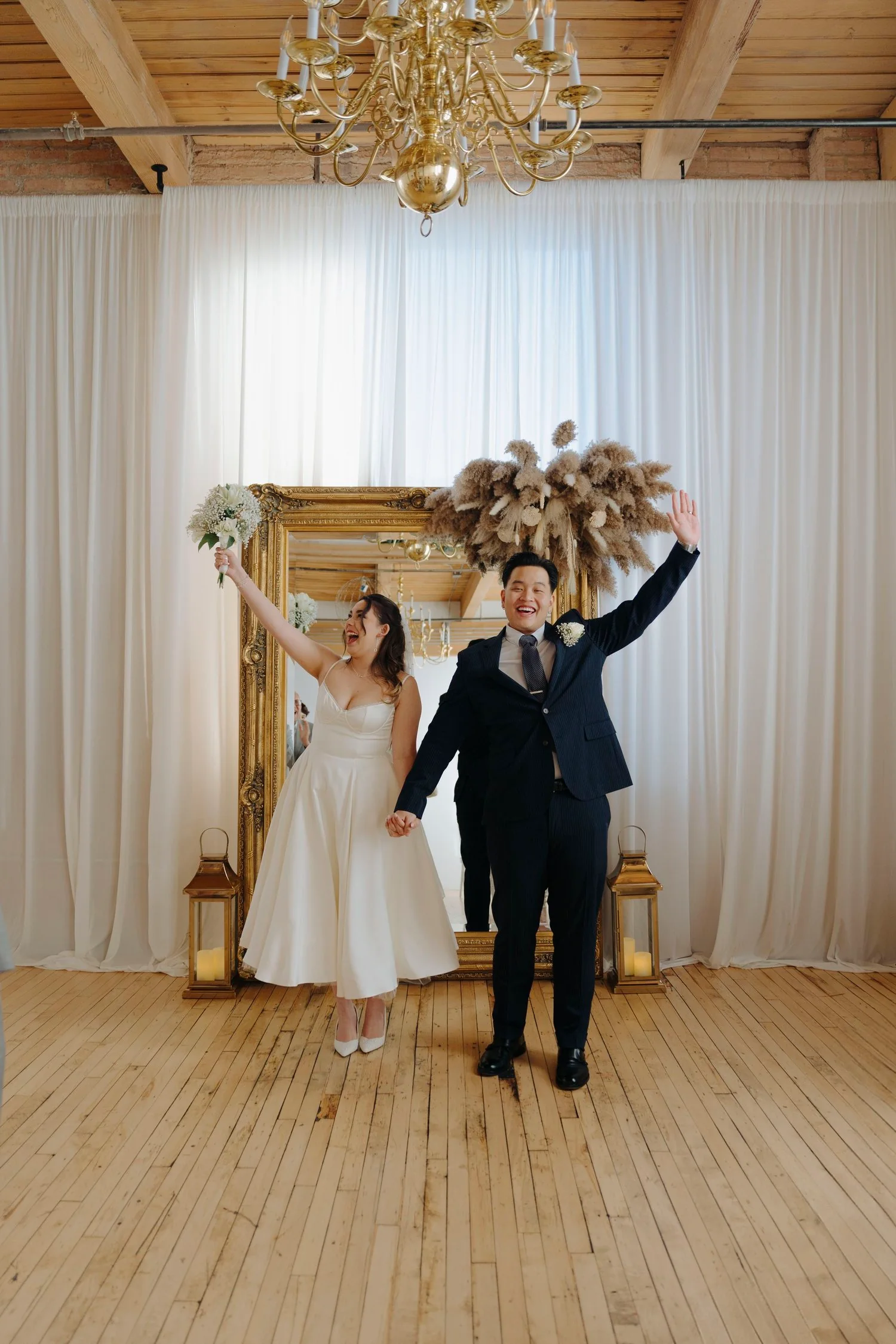 A bride and groom celebrating at their wedding, standing in front of a large mirror with white curtains and wooden floors. The bride is holding a bouquet and smiling, while the groom is raising his hand and holding the bride's hand.
