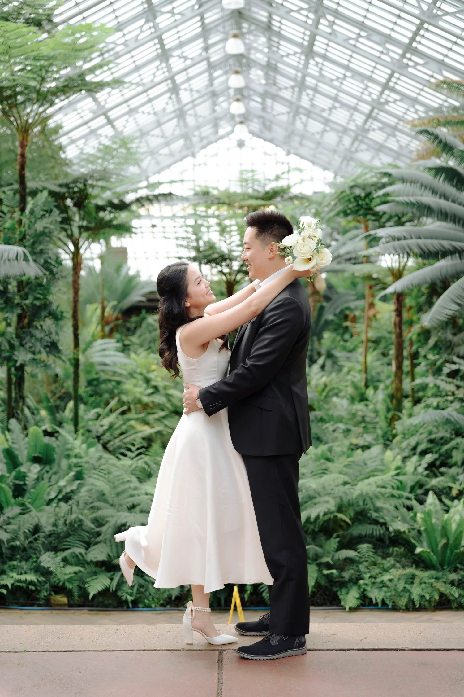 A bride and groom embrace in a greenhouse filled with lush green plants, the bride holding a bouquet of white flowers, both smiling.
