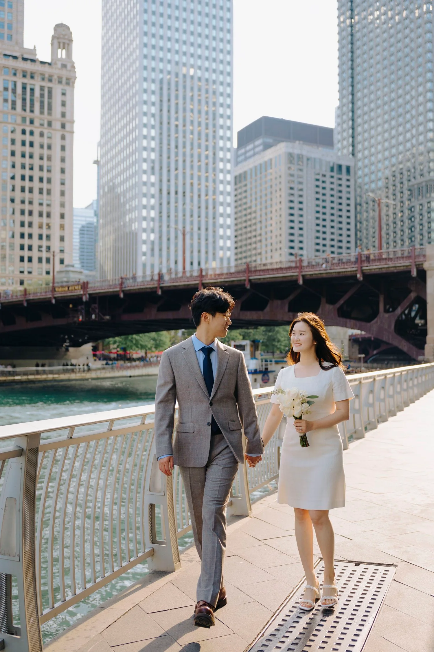 A couple walking hand in hand along a riverwalk in a city with tall skyscrapers in the background, one holding a bouquet of white flowers.
