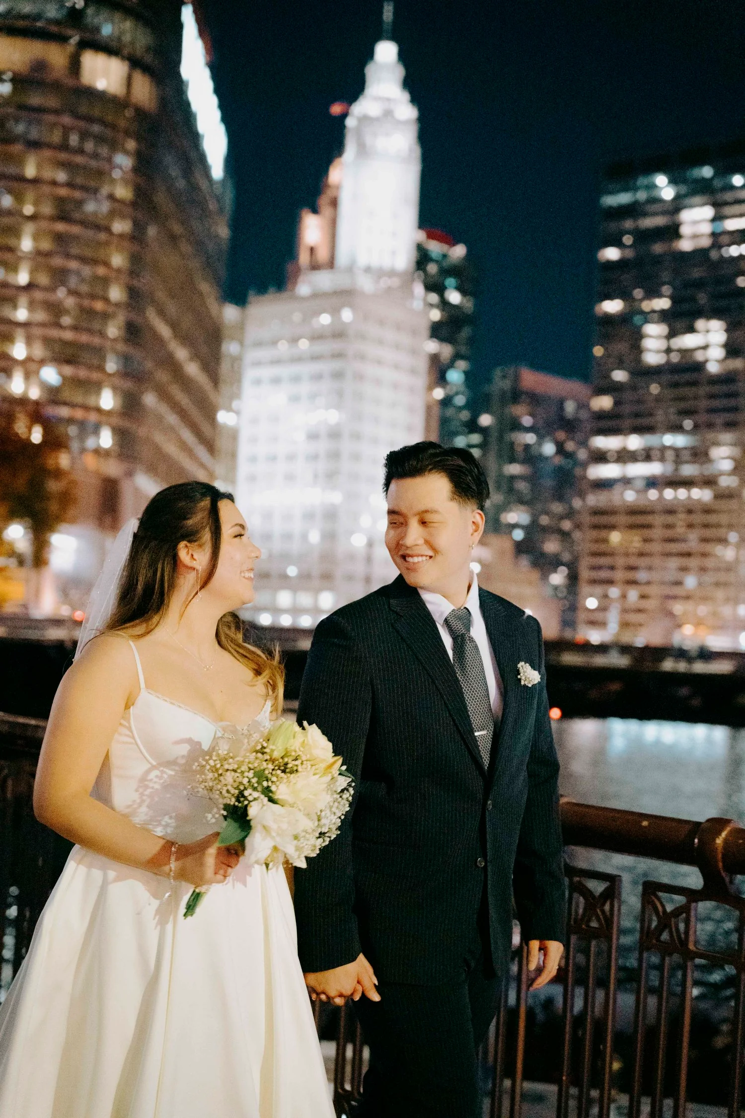 A bride and groom holding hands and smiling at each other outdoors at night with the Empire State Building and city lights in the background.