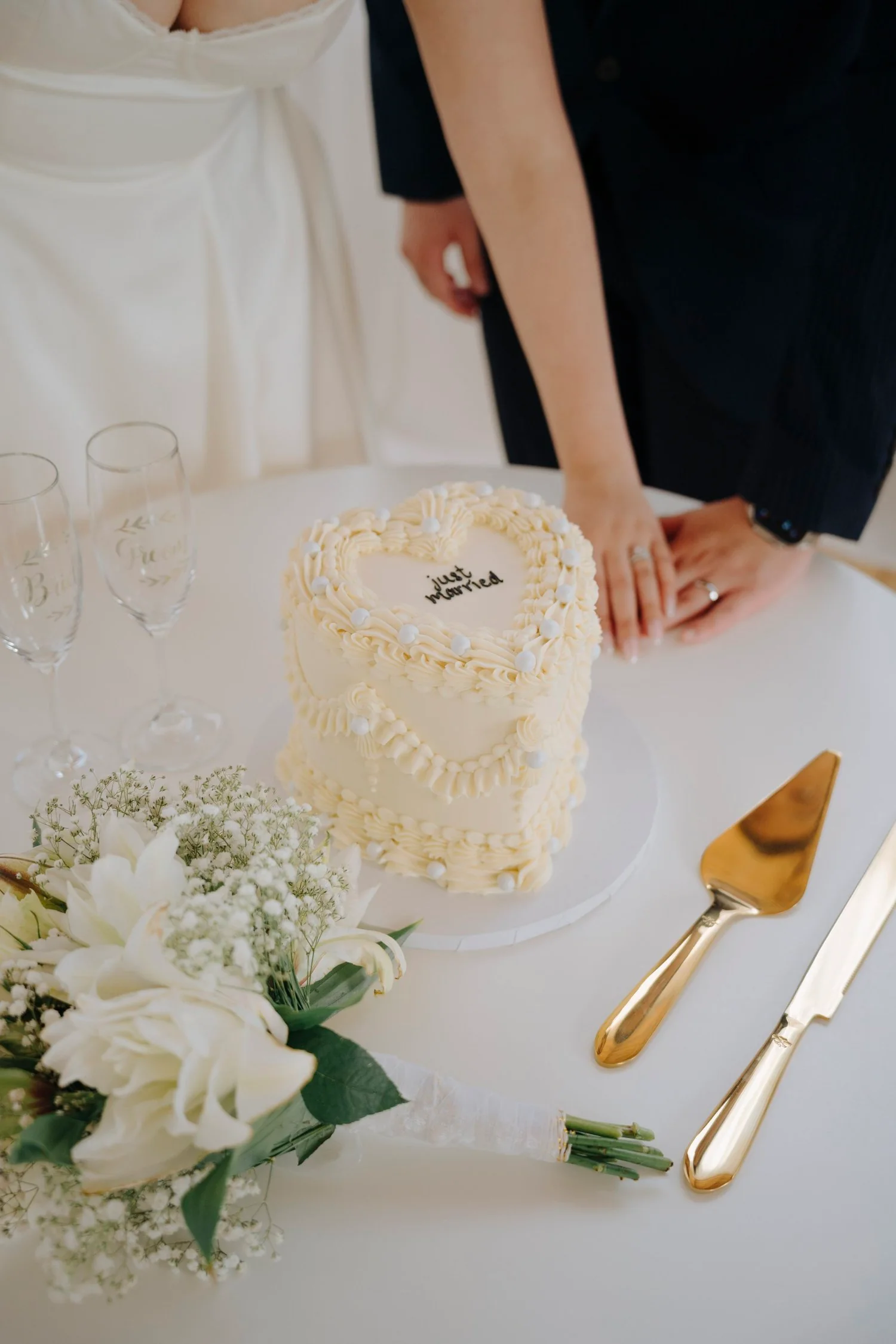 A wedding cake with creamy white icing and the words 'just married' written on top, surrounded by champagne glasses, a bouquet of white flowers, and gold wedding cake utensils on a table.