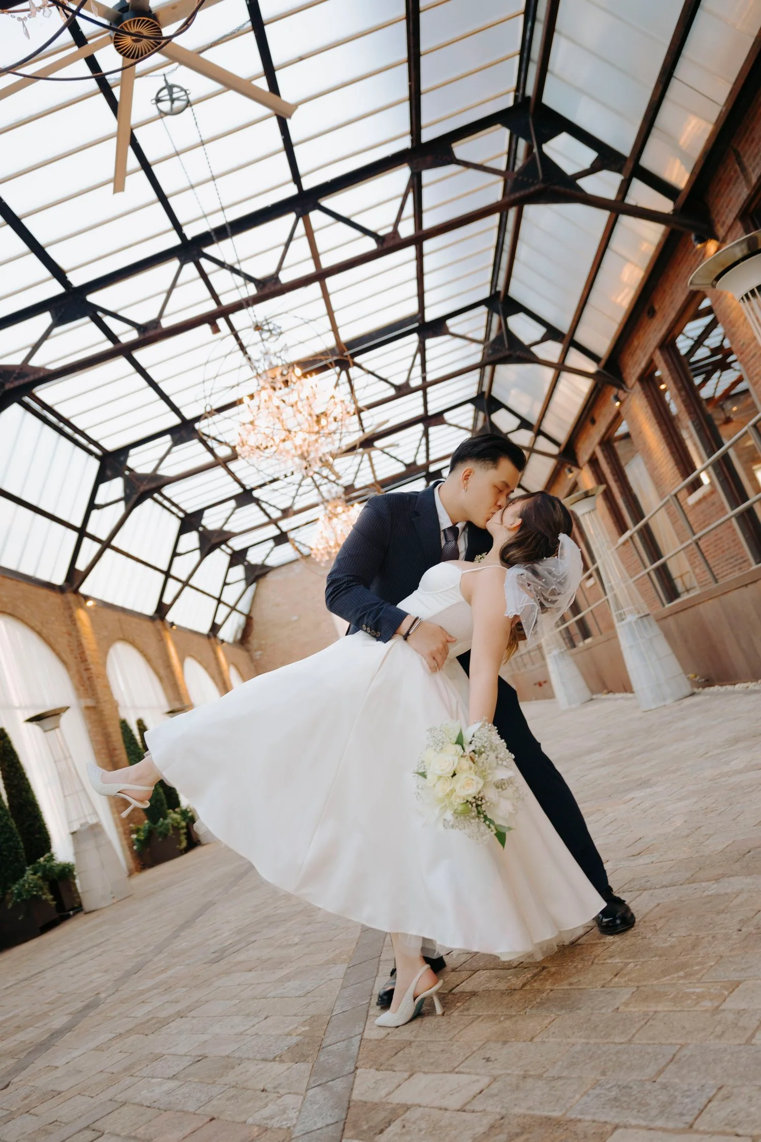 A bride and groom sharing a kiss inside a glass-ceilinged venue, with the groom dipping the bride while she holds a bouquet of white roses.