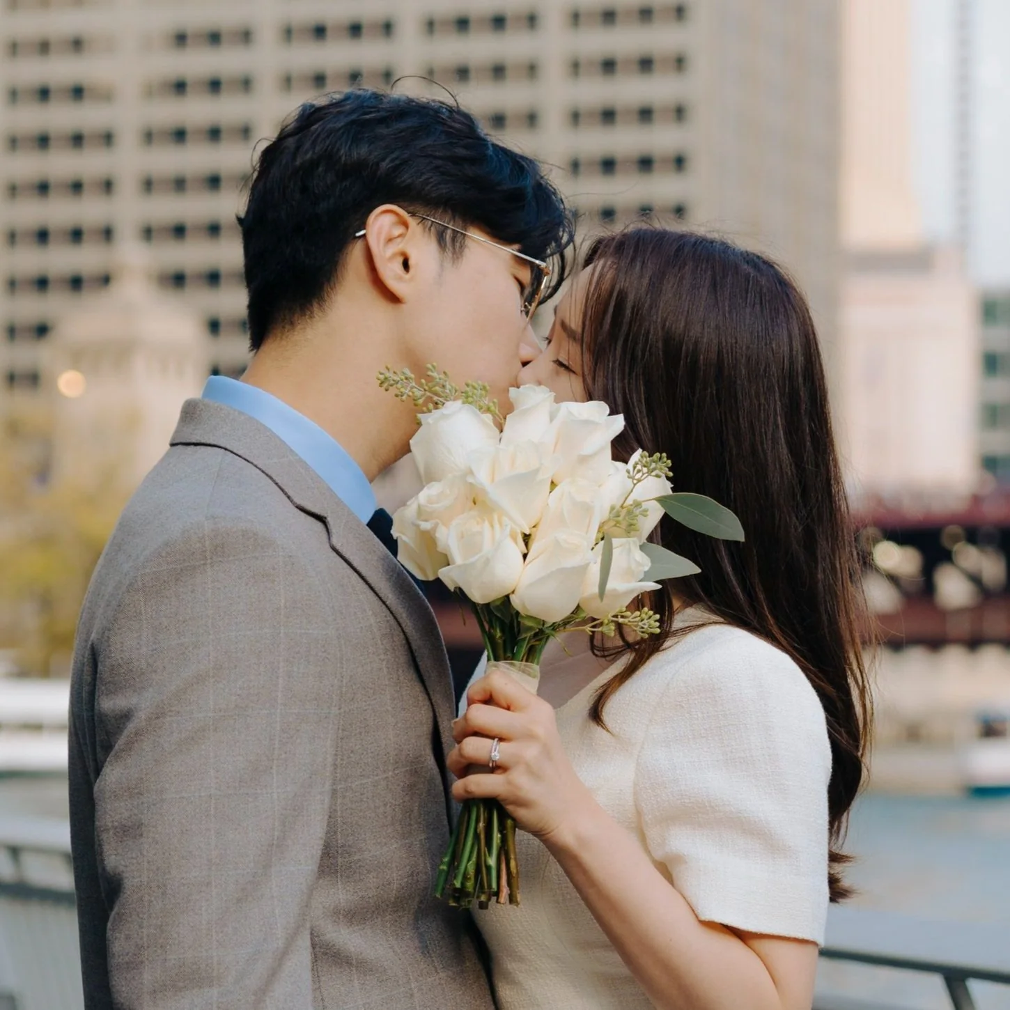 A couple sharing a kiss outdoors with the woman holding a bouquet of white roses.