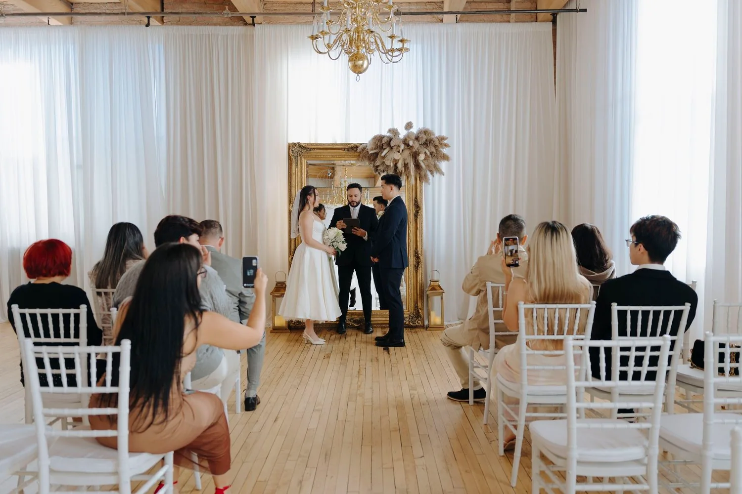 A wedding ceremony taking place indoors with a bride and groom standing before an officiant, surrounded by seated guests capturing the moment on their phones. The venue has large windows, white curtains, and elegant decor including a large gold-framed mirror and a chandelier.