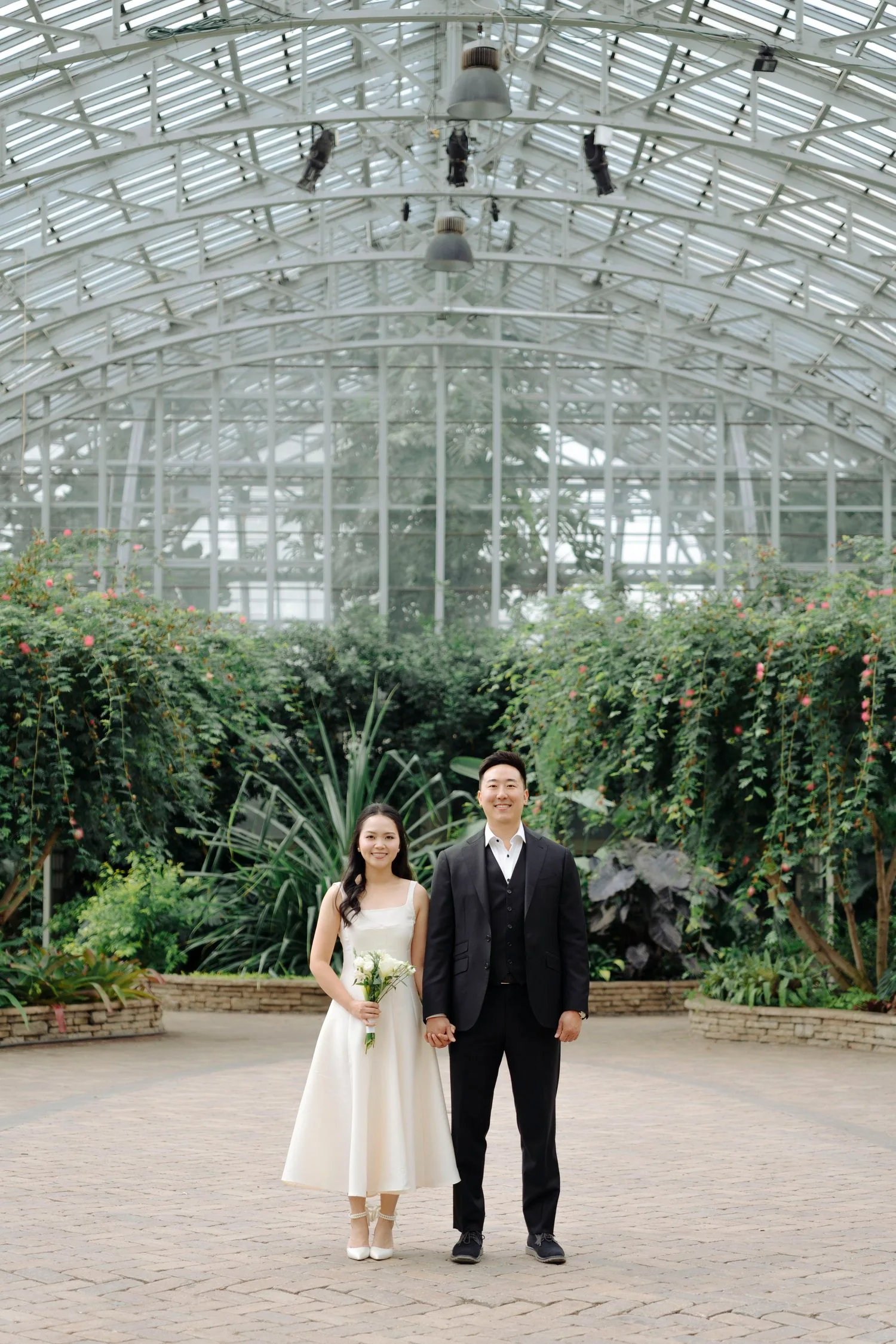 A couple dressed in wedding attire standing inside a greenhouse with lush green plants and flowers, holding hands, with the woman holding a bouquet of white flowers.