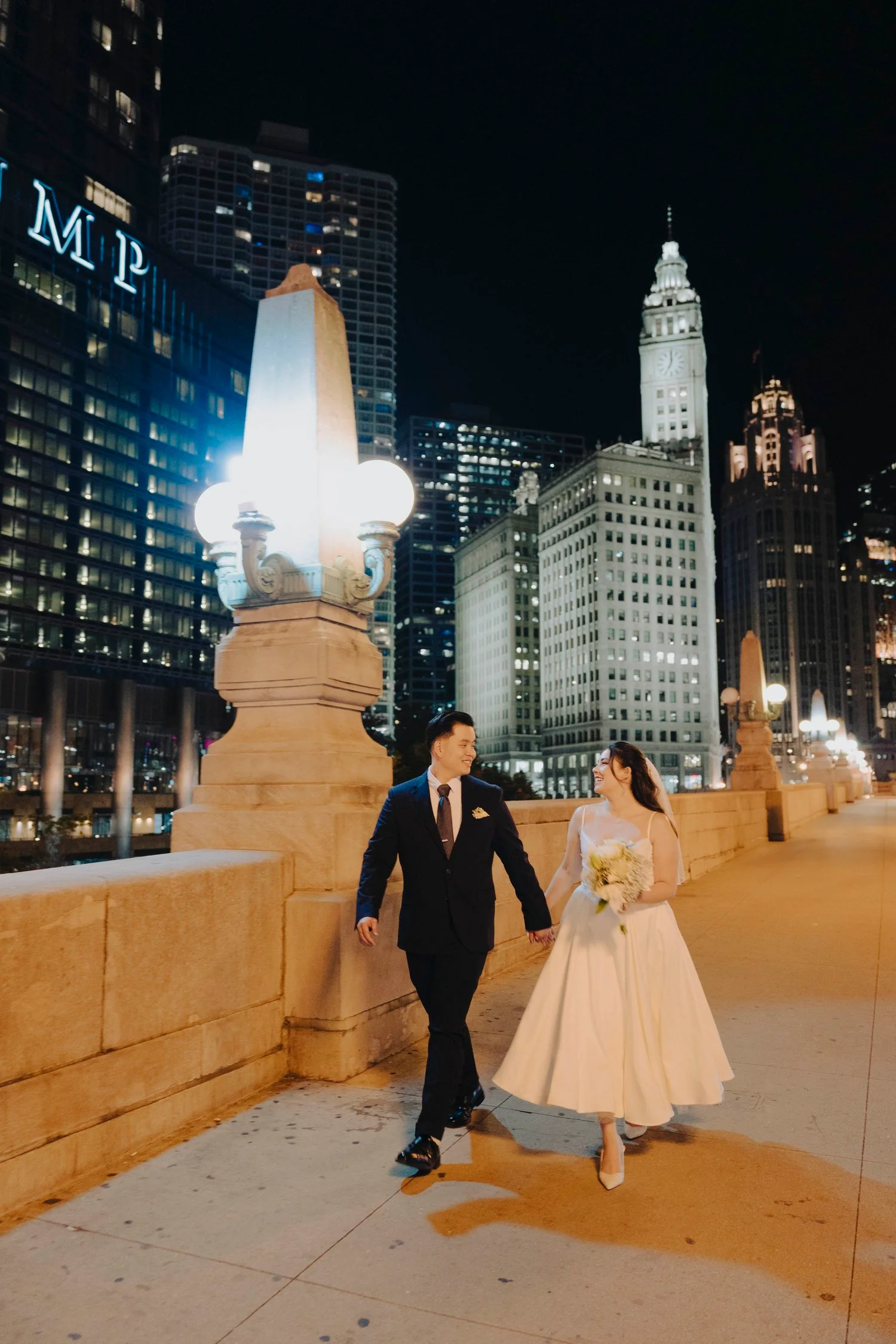 A newlywed couple walking on a city bridge at night, holding hands and smiling, with illuminated skyscrapers and a clock tower in the background.