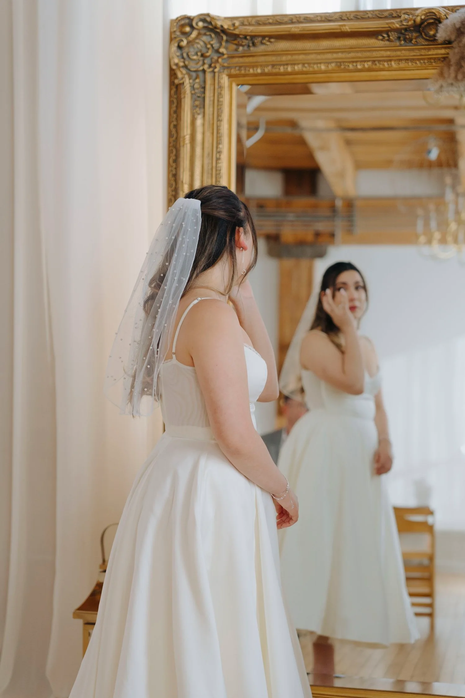 A bride with a veil looking at herself in a mirror, wearing a white wedding dress, with her reflection visible.