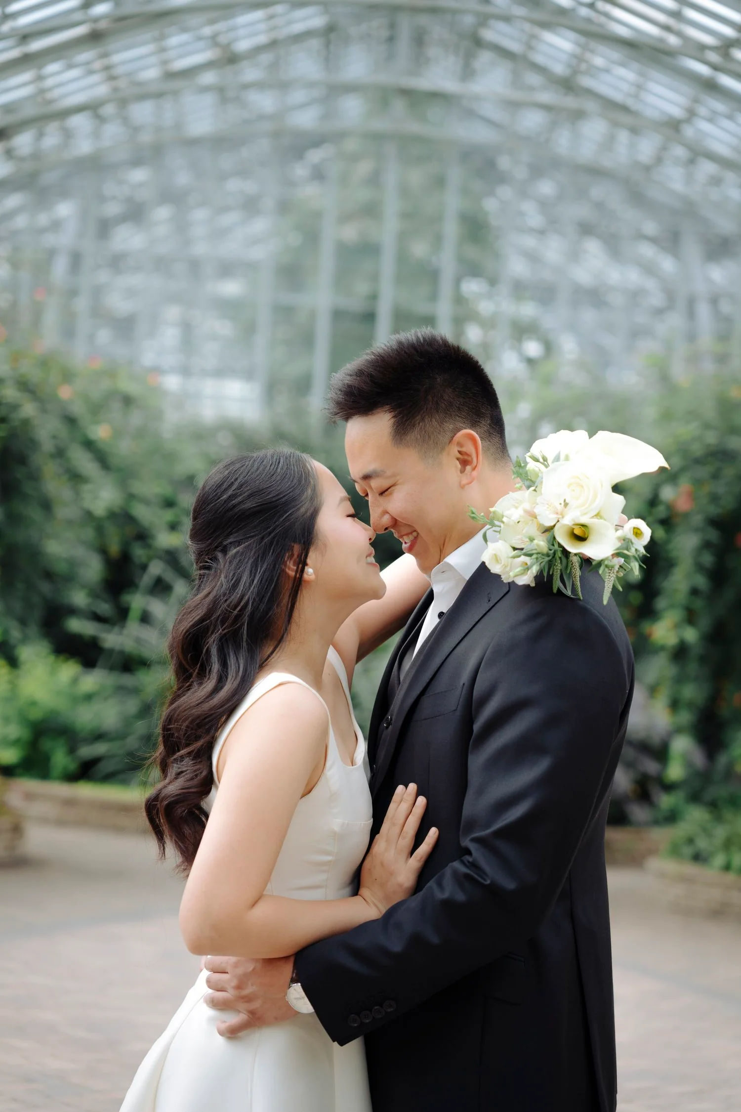 A bride and groom sharing a romantic moment, with faces close and eyes closed, inside a greenhouse.