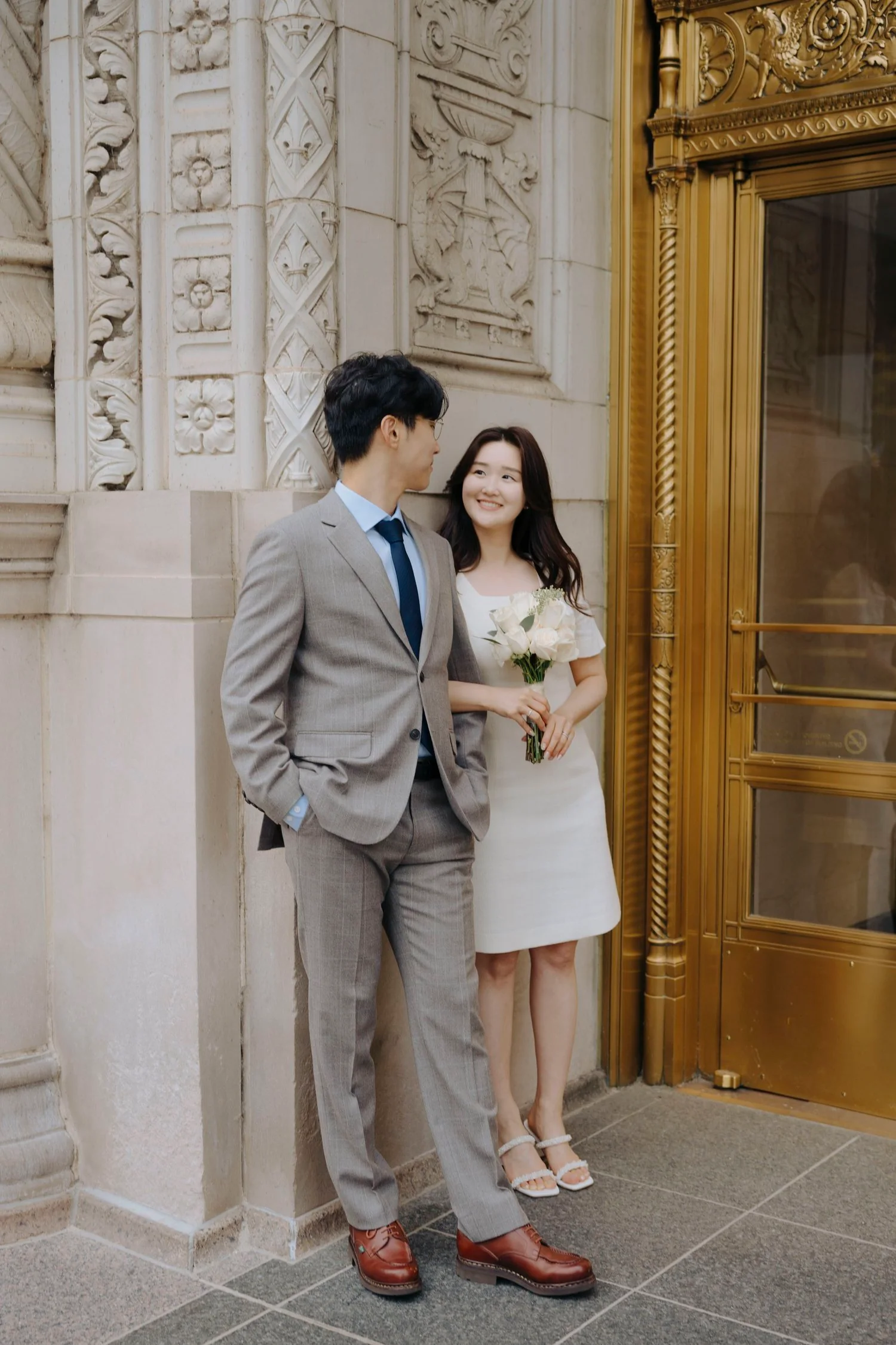 A young woman in a white dress holding a bouquet of white roses stands next to a young man in a gray suit and blue shirt outside a building with ornate stone and gold accents. They are smiling at each other, appearing happy and romantic.